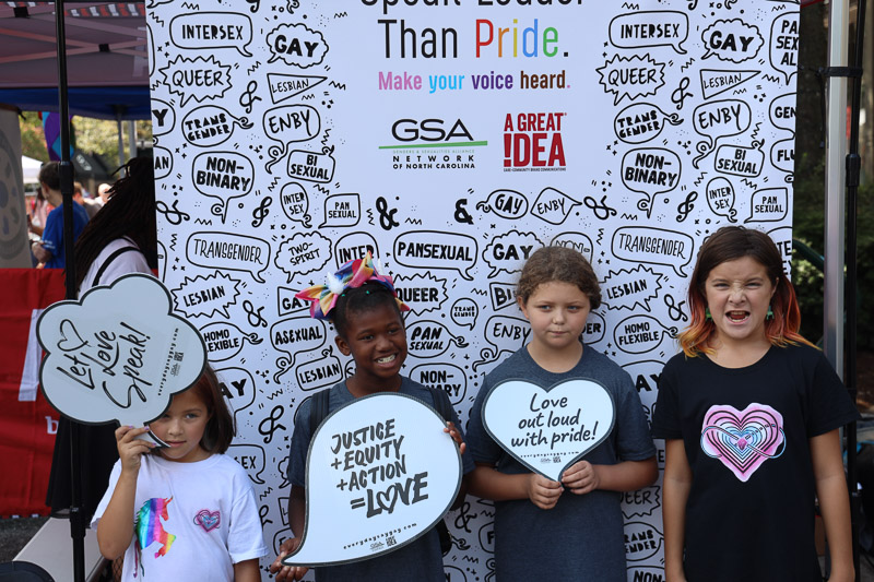 Portrait from Greensboro Pride 2022 with attendees in front of wall saying 'Actions Speak Louder Than Pride. Make Your Voice Heard." from Genders and Sexualities Alliance Network of North Carolina and A Great Idea, a Care and Community Brand Communications Agency. Photo courtesy of A Great Idea (https://weareagi.com)