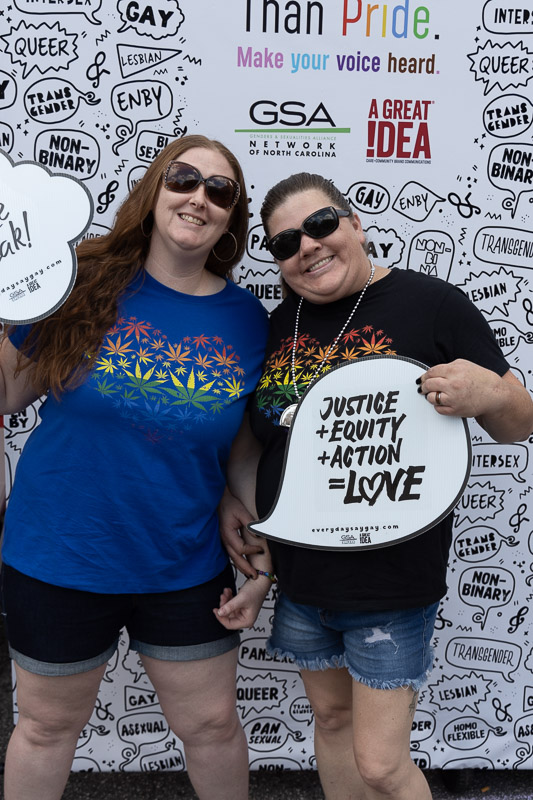 Portrait from Greensboro Pride 2022 with attendees in front of wall saying 'Actions Speak Louder Than Pride. Make Your Voice Heard." from Genders and Sexualities Alliance Network of North Carolina and A Great Idea, a Care and Community Brand Communications Agency. Photo courtesy of A Great Idea (https://weareagi.com)