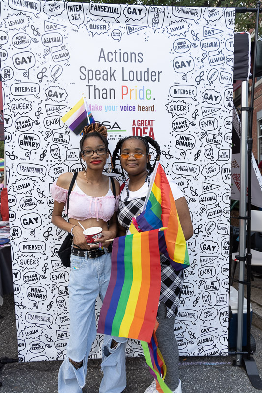 Portrait from Greensboro Pride 2022 with attendees in front of wall saying 'Actions Speak Louder Than Pride. Make Your Voice Heard." from Genders and Sexualities Alliance Network of North Carolina and A Great Idea, a Care and Community Brand Communications Agency